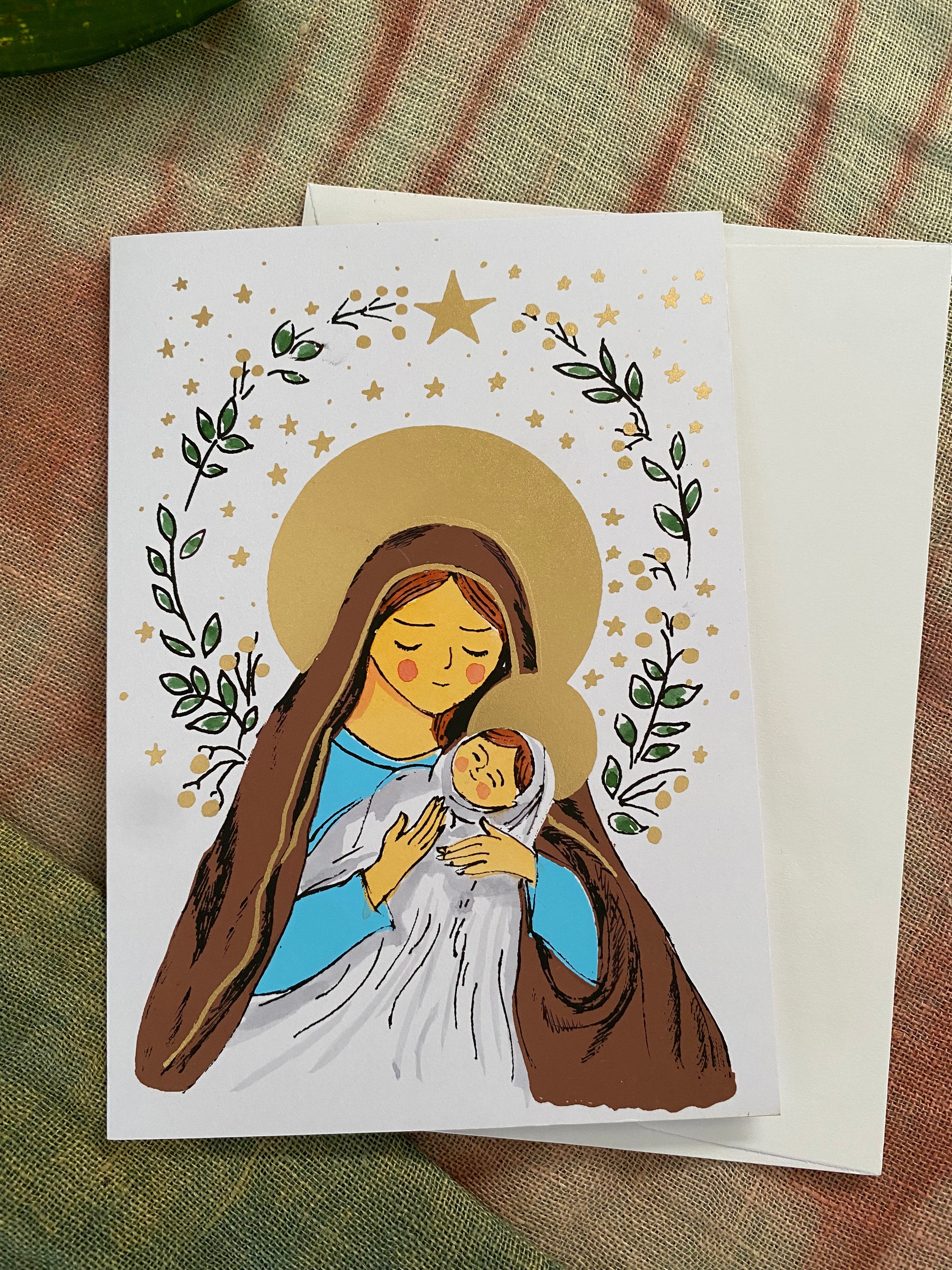 Christmas Card with illustration of a woman , Mary, holding a baby, surrounded by stars and leaves on a textured fabric background.