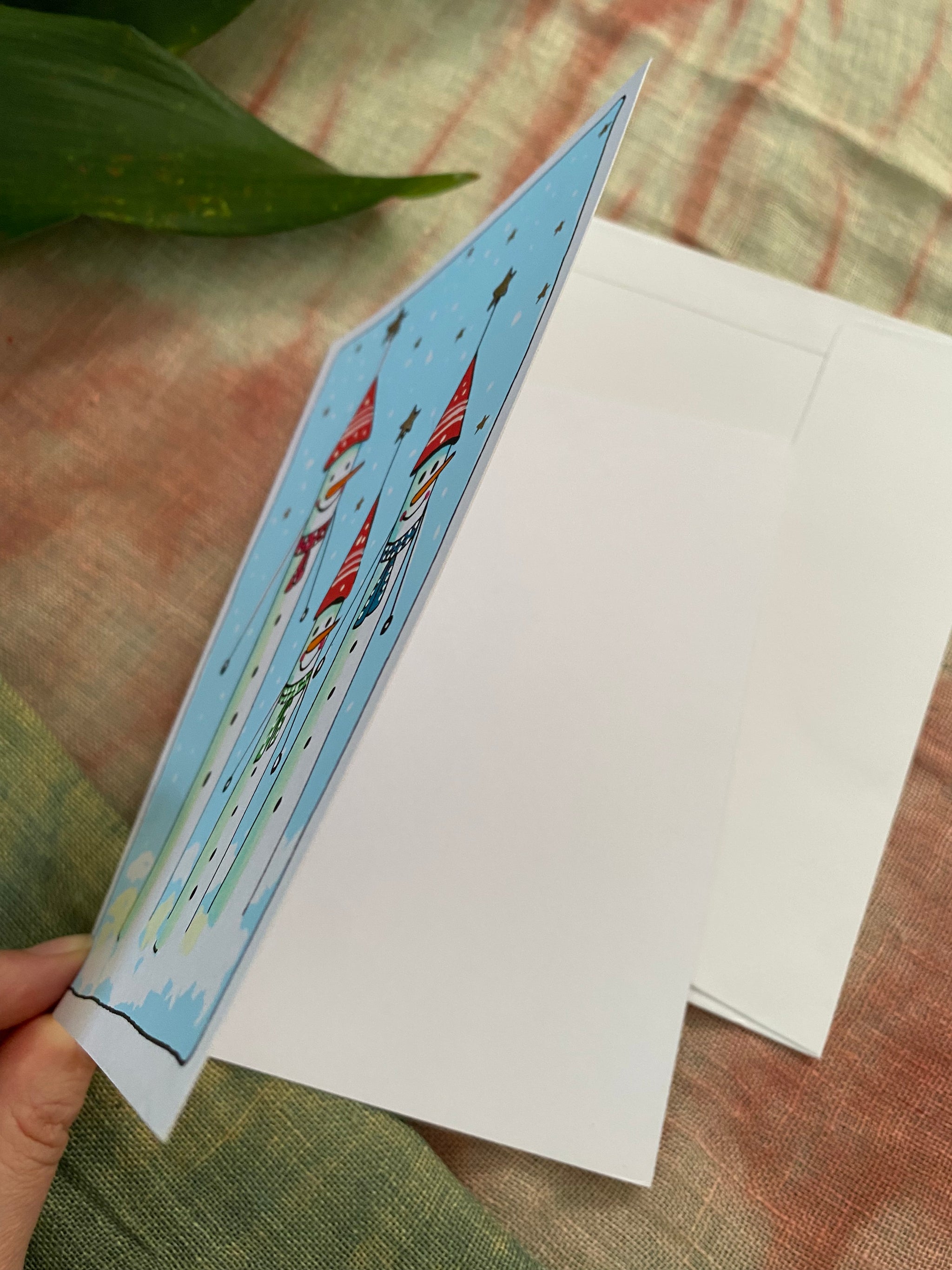 Christmas card with snow people illustration held over a white envelope on a textured surface.