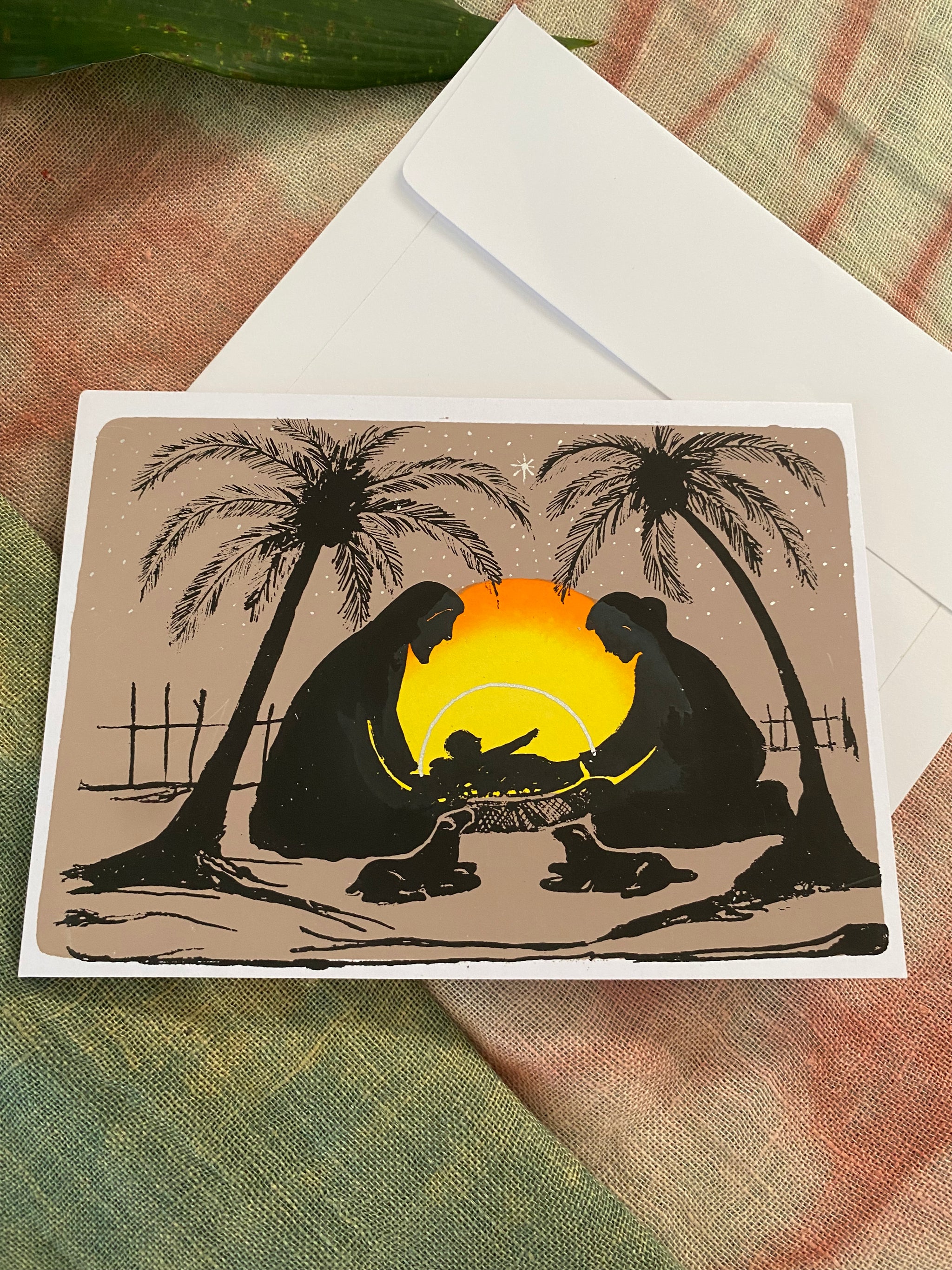Christmas card landscape-format card showing Mary, Joseph and baby Jesus in silhouette beneath a glowing sunset. Palm trees, sheep and the setting sun frame the holy family,