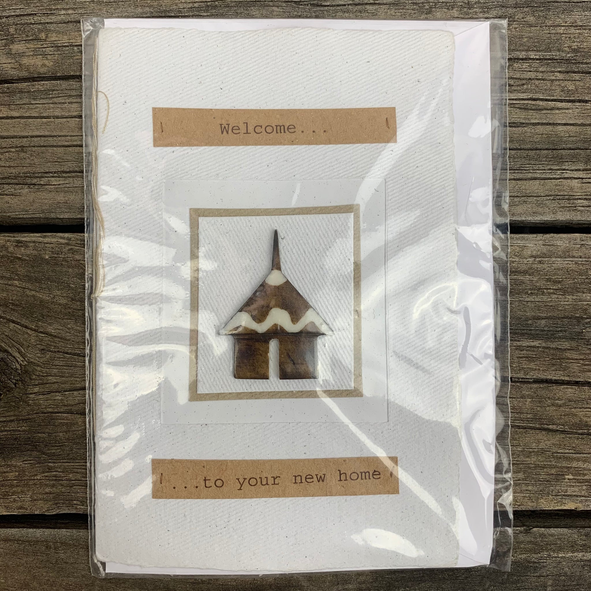 greeting card displaying message - 'welcome to your new home'. The card is white with text bordered by brown recycled paper. In the centre of the card there is a small tribal house that protrudes from the surface