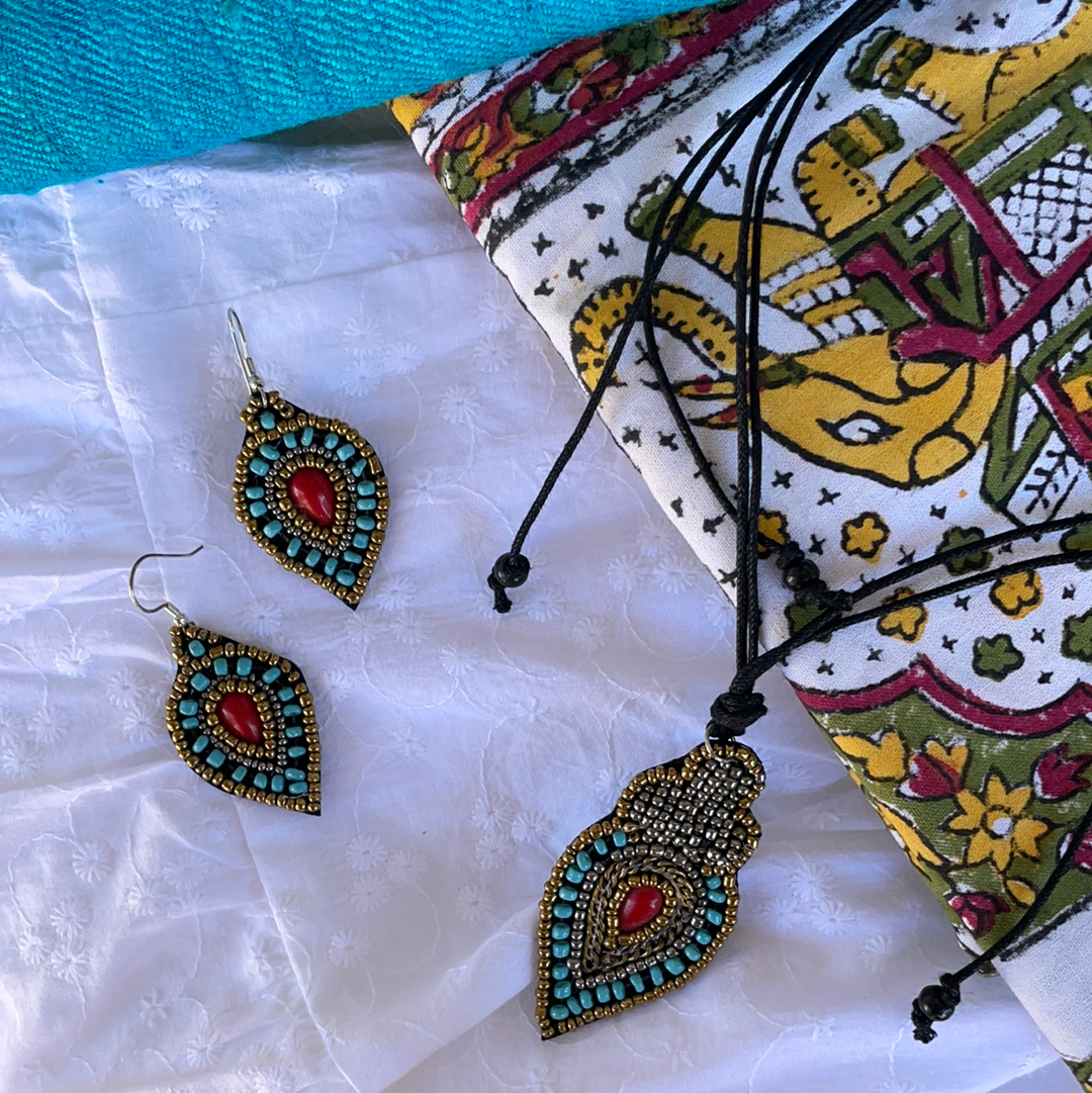 a red center tear drop shaped bead with small gold beads surrounding the red bead, followed by larger blue beads and another layer of small gold beads hand sown onto a soft felt backing and make into earrings and necklace with a corded tie.