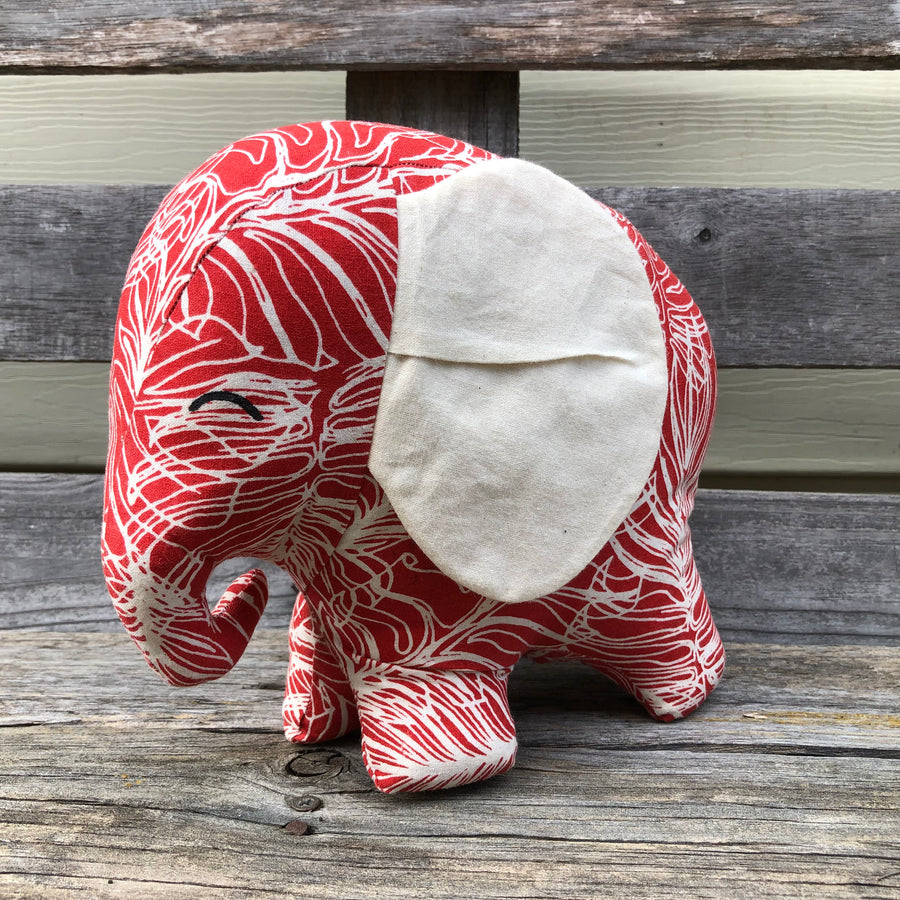 Red elephant with white screeen printed leaf outlines and big white fabric ears sitting on a wooden bench facing to the side