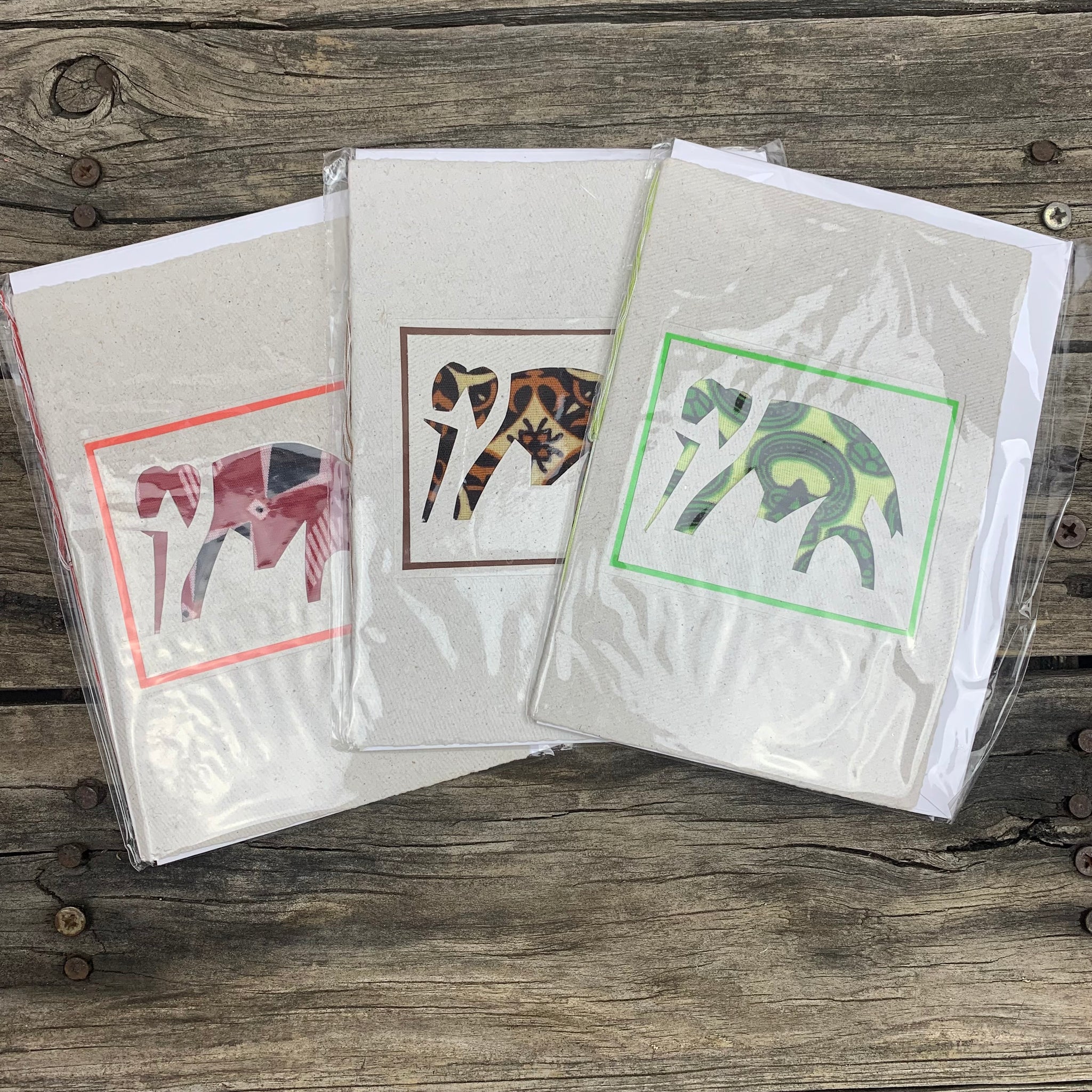 White handmade paper greeting card displaying an elephant in the centre. There are 3 differentiating designs, a red elephant, a brown one, and a green one - all bordered by their respective colours 