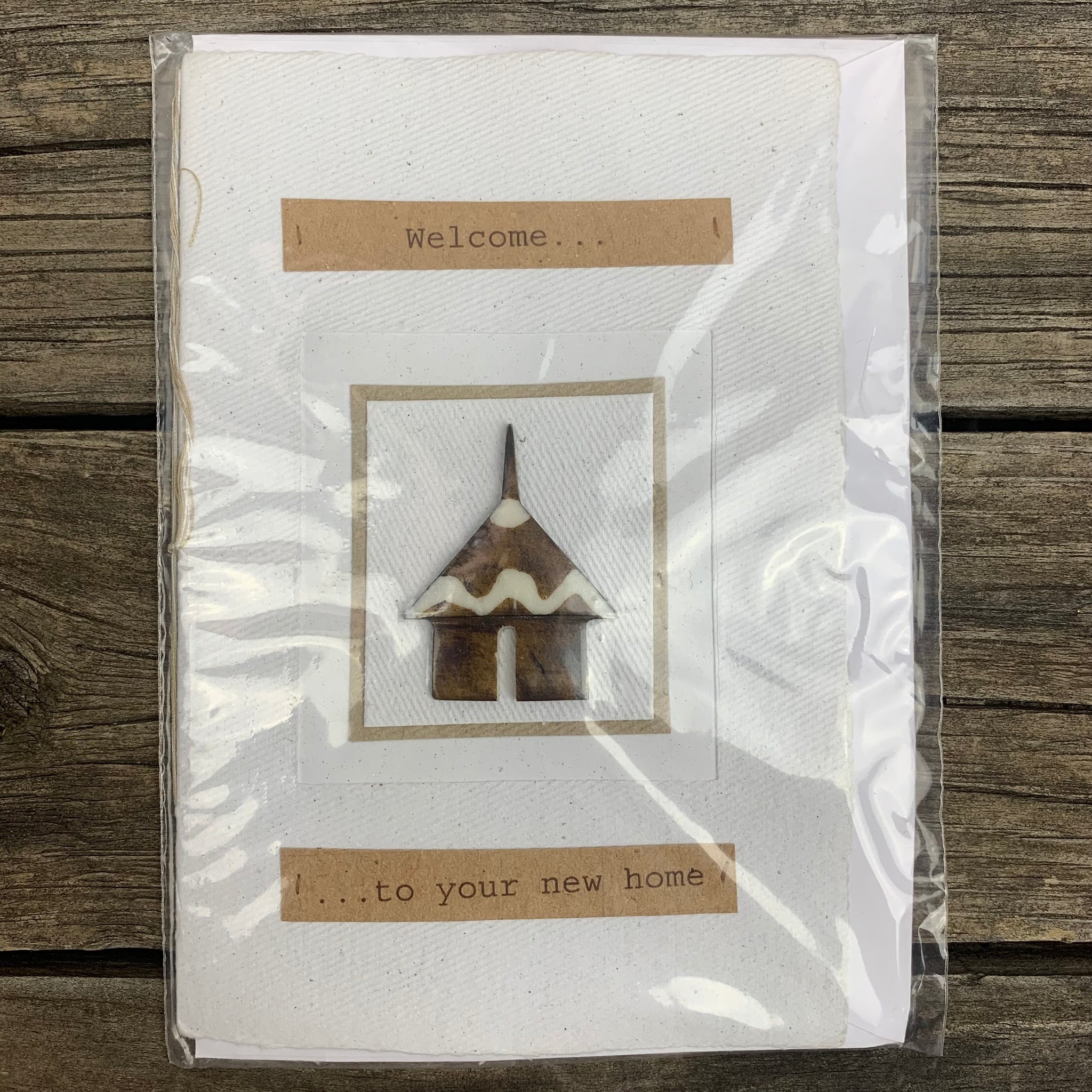 greeting card displaying message - 'welcome to your new home'. The card is white with text bordered by brown recycled paper. In the centre of the card there is a small tribal house that protrudes from the surface 