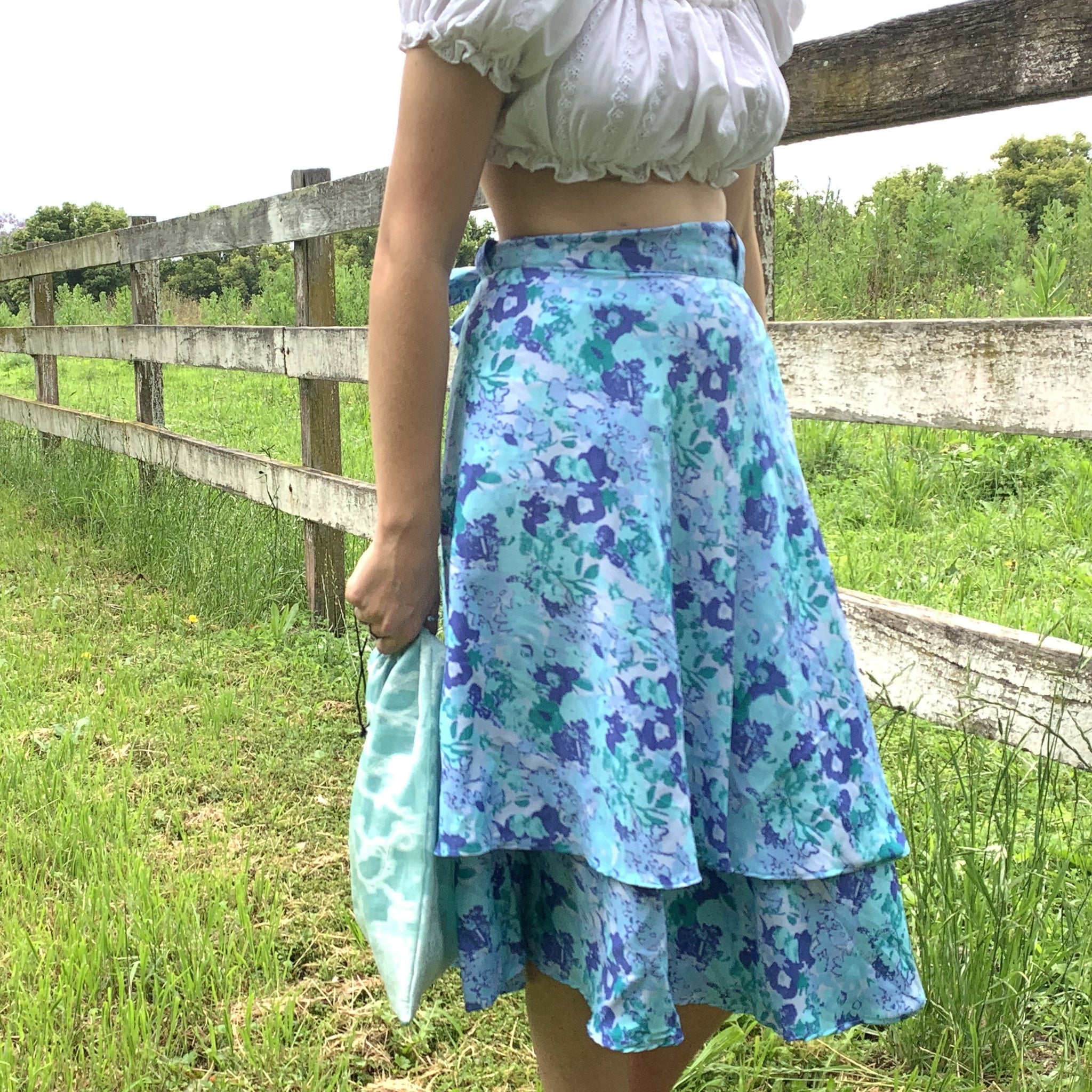 Lydia is wearing a blue double layered wrap skirt with a white crop. The blue skirt features lots of different shades of blue in a subtle flower pattern. She is standing infront of an old white fence in a field of grass. 