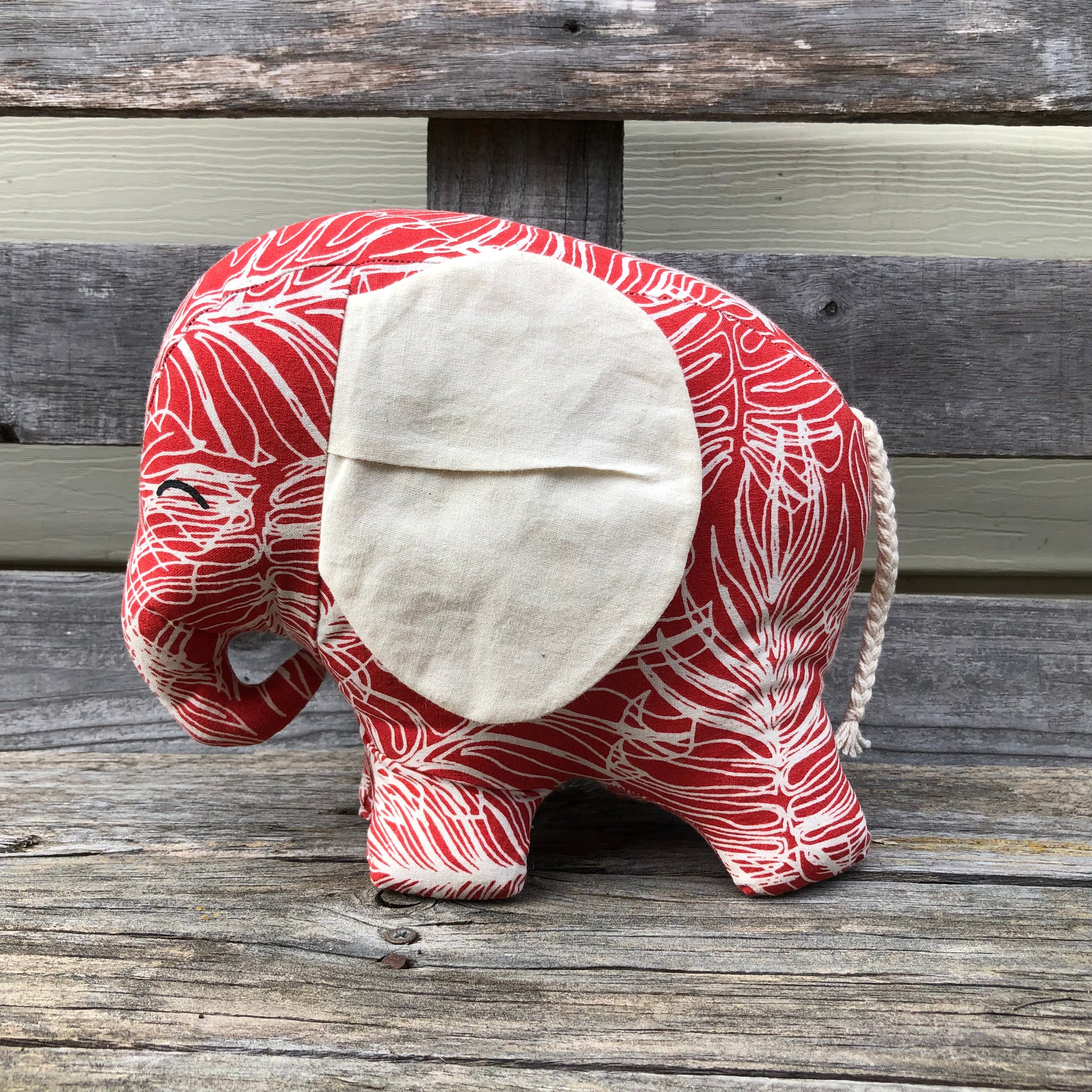 Red elephant with white screeen printed leaf outlines and big white fabric ears sitting on a wooden bench facing to the side